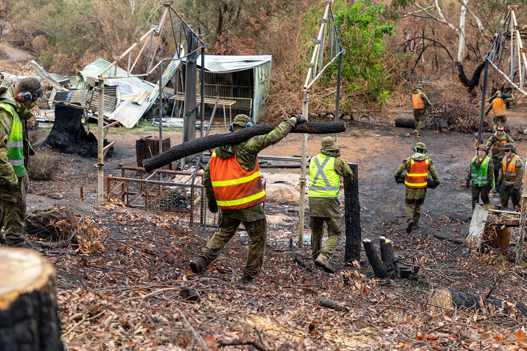 Australian Defence Force (ADF) reservists wearing high-visibility vests over their camouflage uniform. They are removing and carrying branches, logs and stumps away from the bushforest. There is the burnt remains of a large shed or house and a small shed.