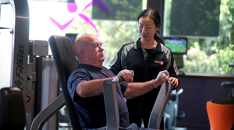 Young woman watches over elderly man on weights machine in gym