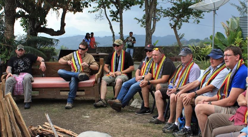 Eight or so men outside sitting in semi-circle listening to someone with tropical scenery in background