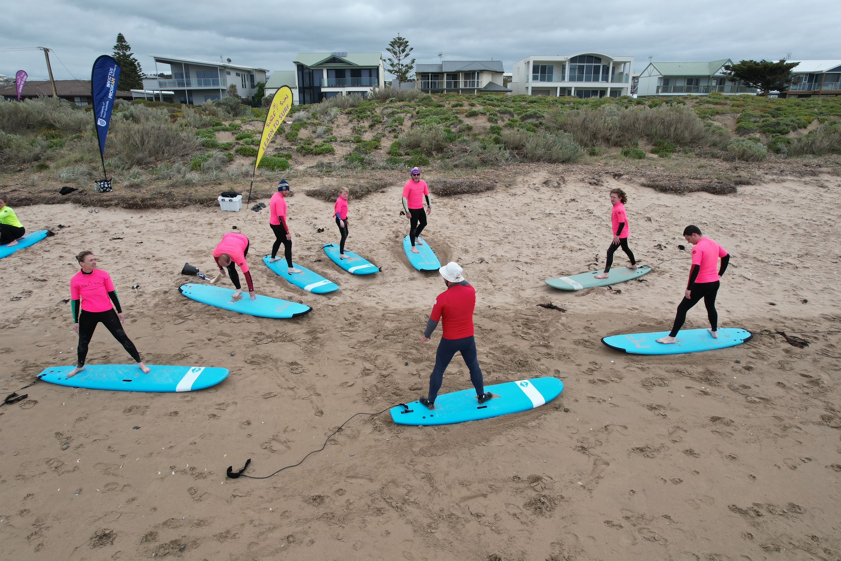 A group of 8 people are wearing pink and black wetsuits, standing on surfboards on the sand at Middleton Beach, South Australia