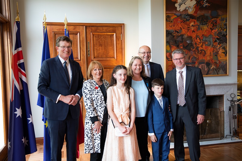 Three men, two women and two children pose for camera in large nicely decorated room, next to Australian flag