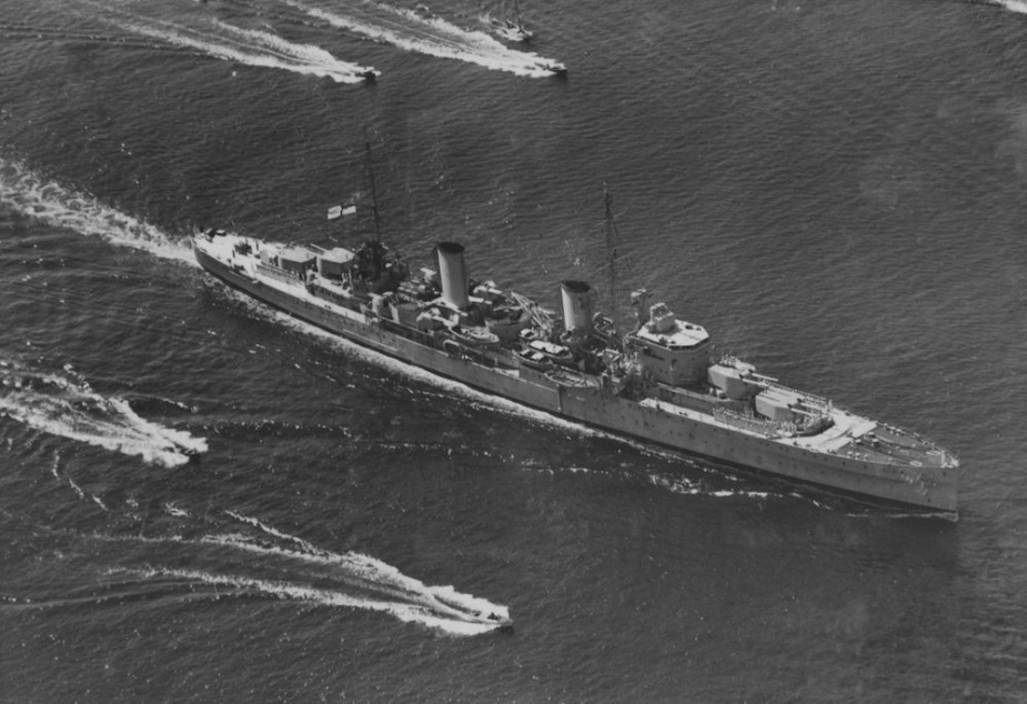 Aerial photo of Second World War cruiser on calm water surrounded by smaller boats