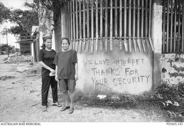 Two East Timorese women stand next to wall that the words 'We love interfet thanks for your security' written on it