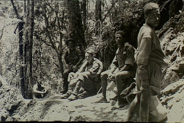 Black and white photo of Australian soldiers sitting next to Papuan bearers