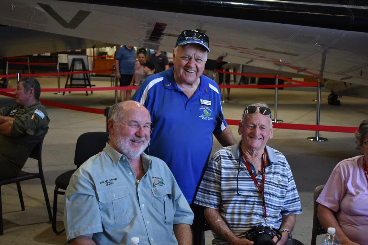Three older men smiling, seated next to aircraft in hangar