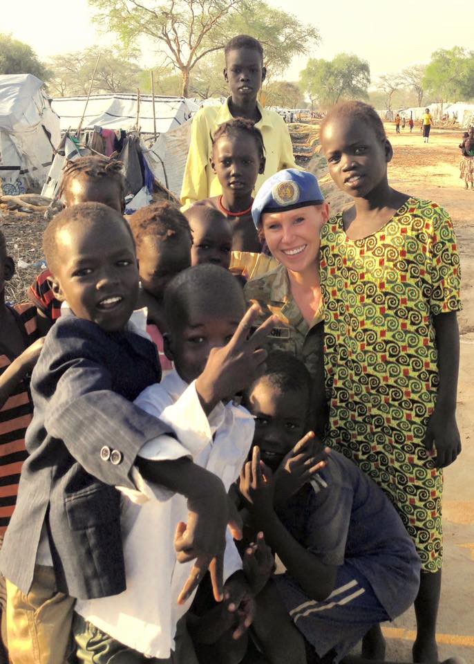 Claire Pearson in uniform kneels, surrounded by children with tents in the background.