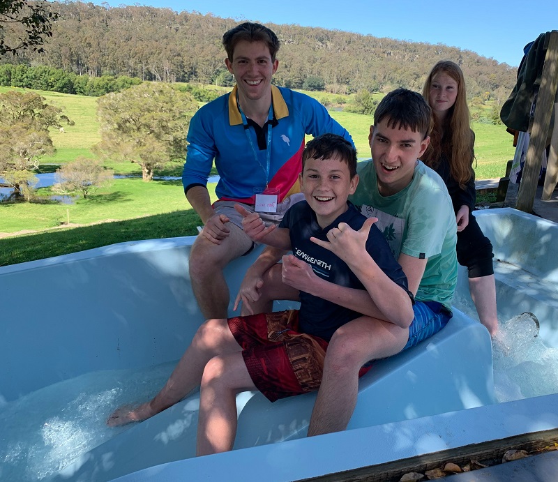 A Kookaburra Kids team members sits with three kids at the top of a water slide. The background shows grass and trees.