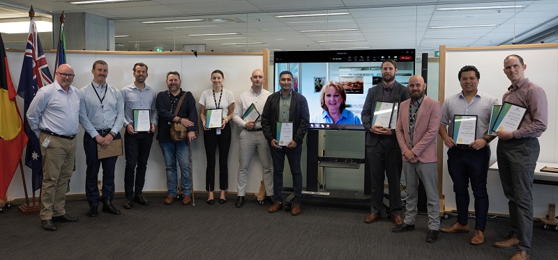 A group of people involved with the pilot program stand in a semi-circle holding certificates.