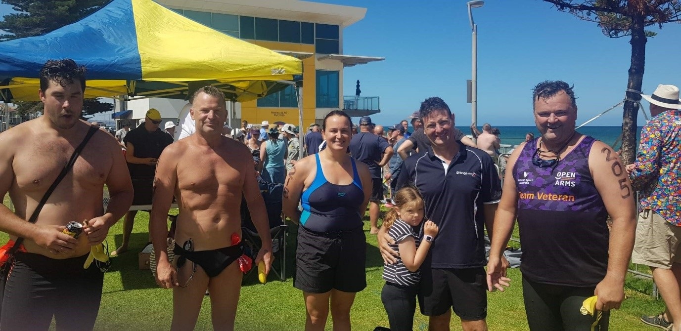 A group of people in swimming costumes stand in front of a blue and yellow gazebo.