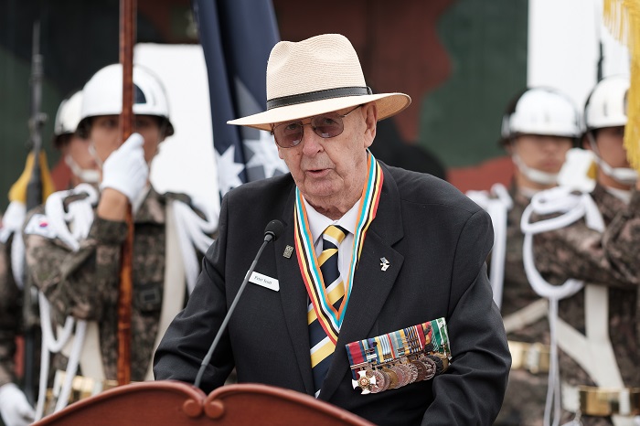 Peter, wearing his medals, is delivering a reading at a lecturn with a microphone in front of him.