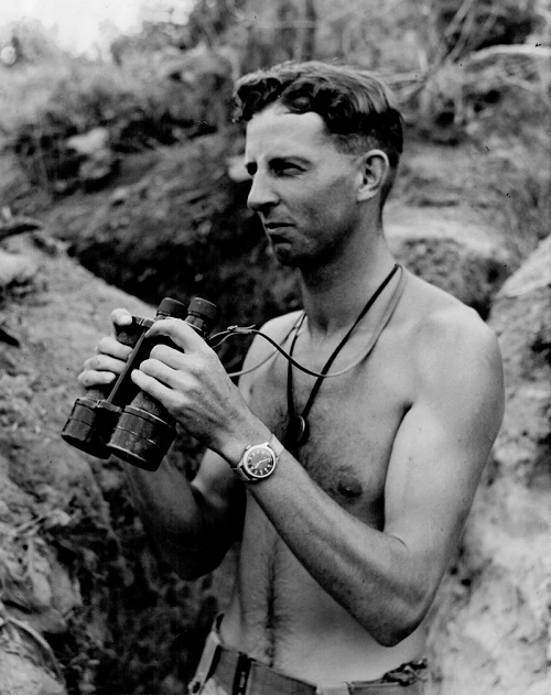 Peter stands in a trench without a shirt on, holding binoculars.