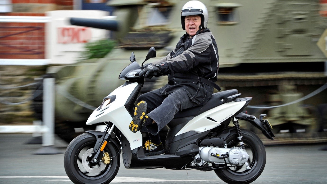 An older man grinning as he rides a motor scooter