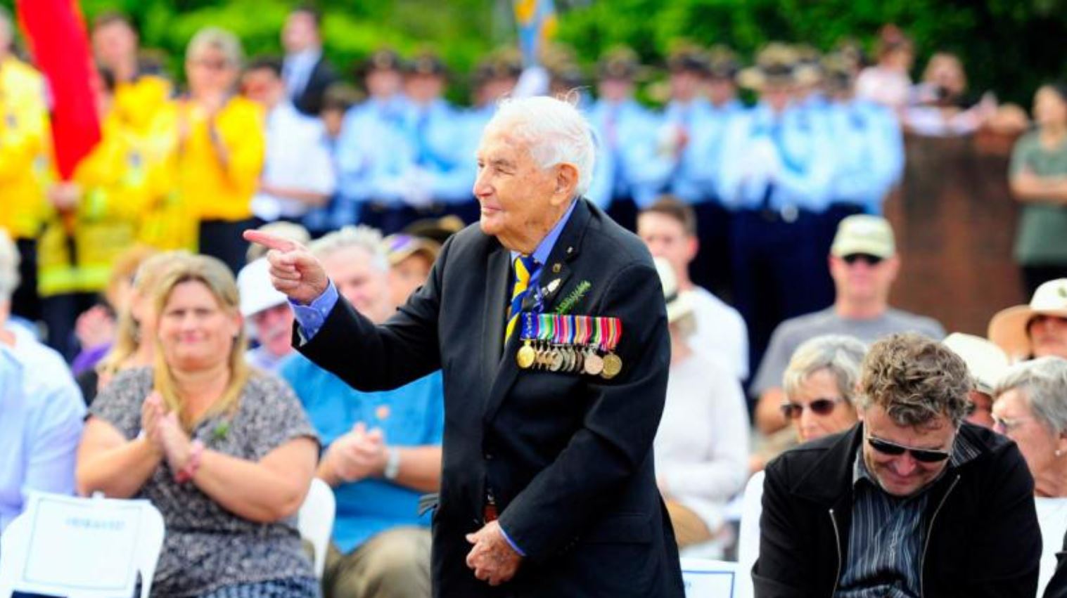 Older man wearing medals standing up in front of applauding crowd