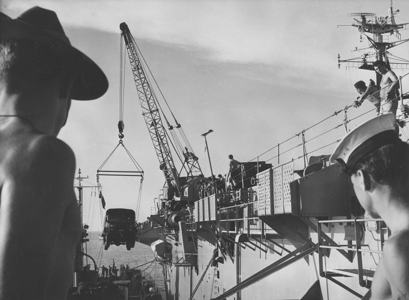 A soldier and a sailor watch a jeep being loaded or unloaded onto an aircraft carrier