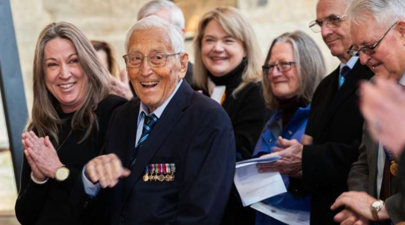 Elderly man wearing medals and smiling, surrounded by others outside a church