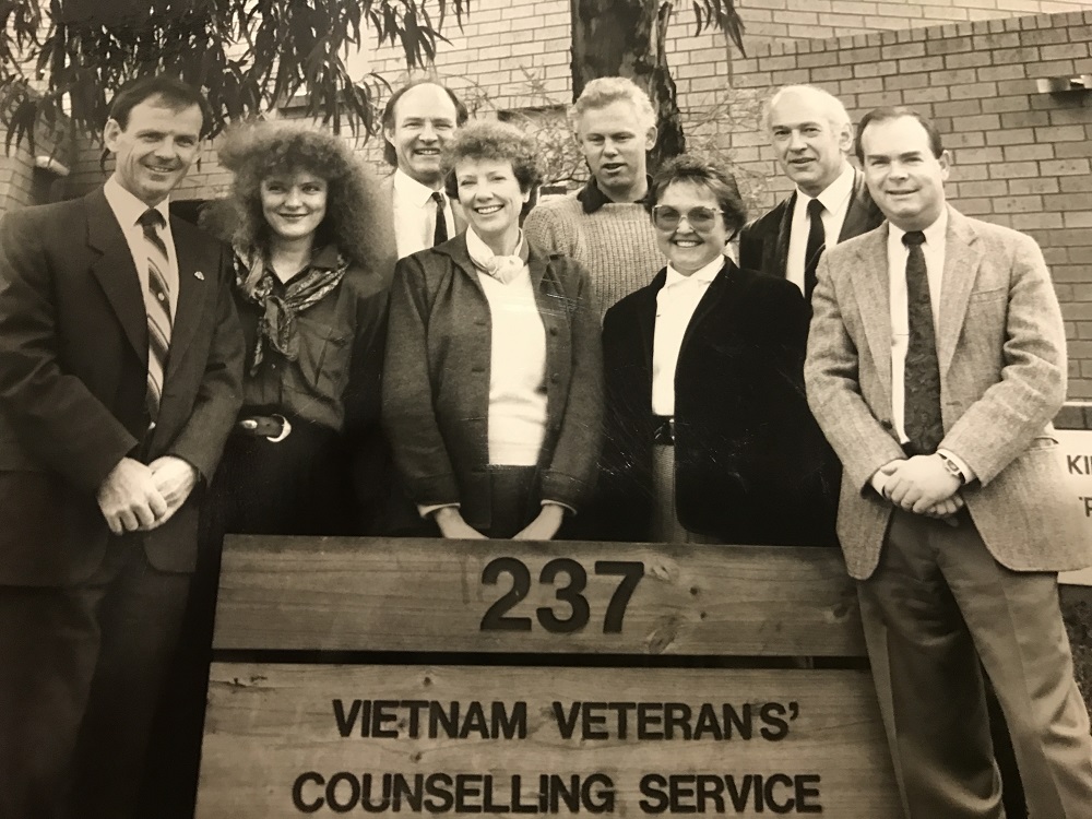 Black and white photo of eight men and women posing next to sign that says Vietnam Veterans' Counselling Service