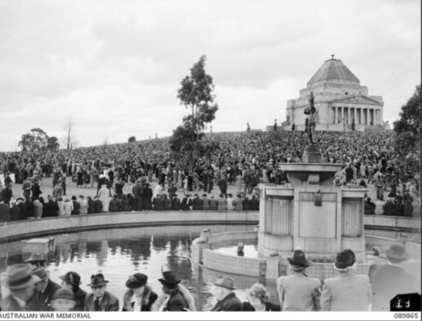 Black and white photo of thousands of people. A fountain is in the foreground, and in the background is a large building with a pyramidal roof.