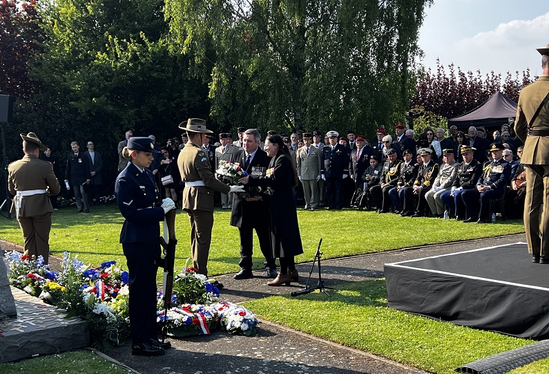 Man and woman present wreath to soldier at cenotaph with crowd of people seated in background