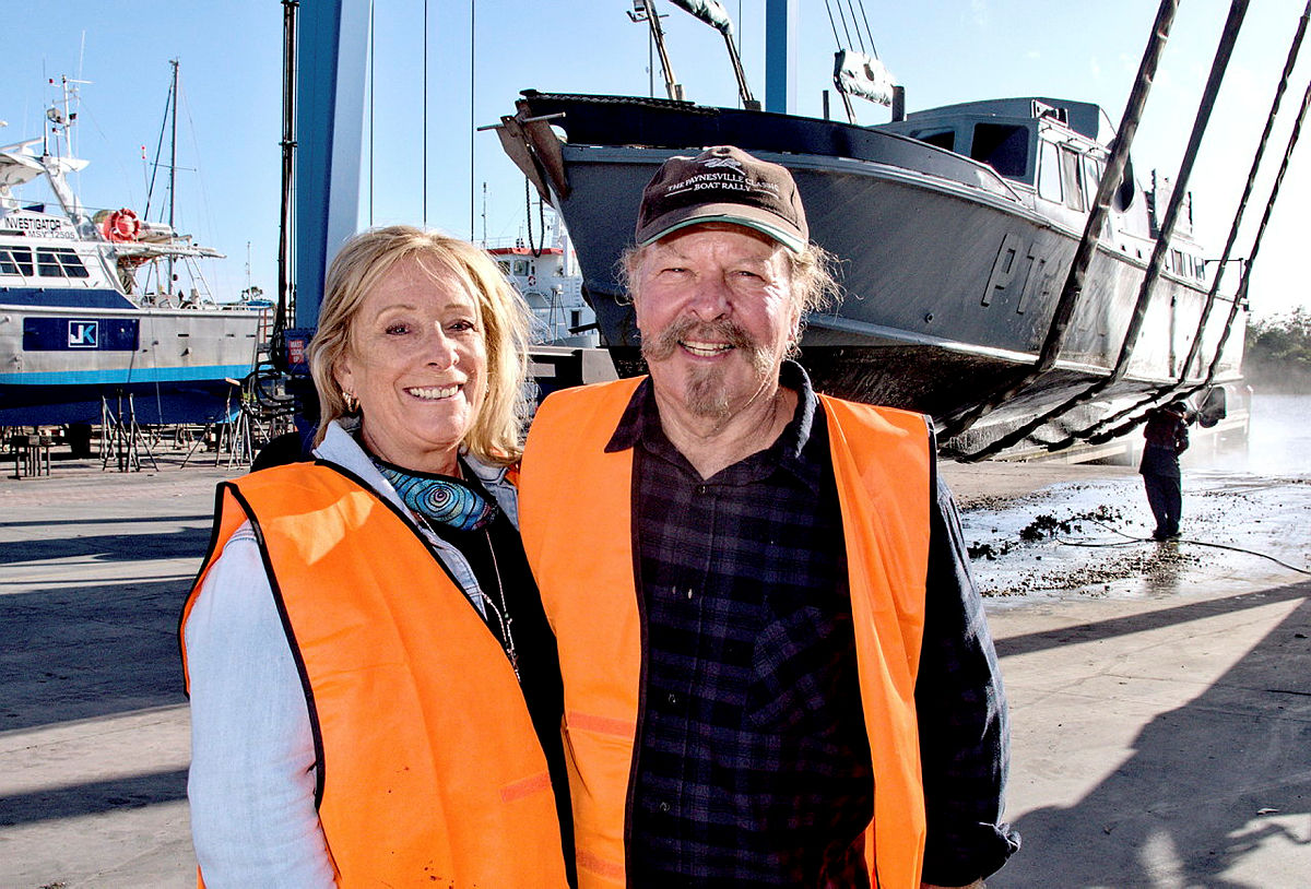 Couple pose for camera in front of grey boat suspended in harness