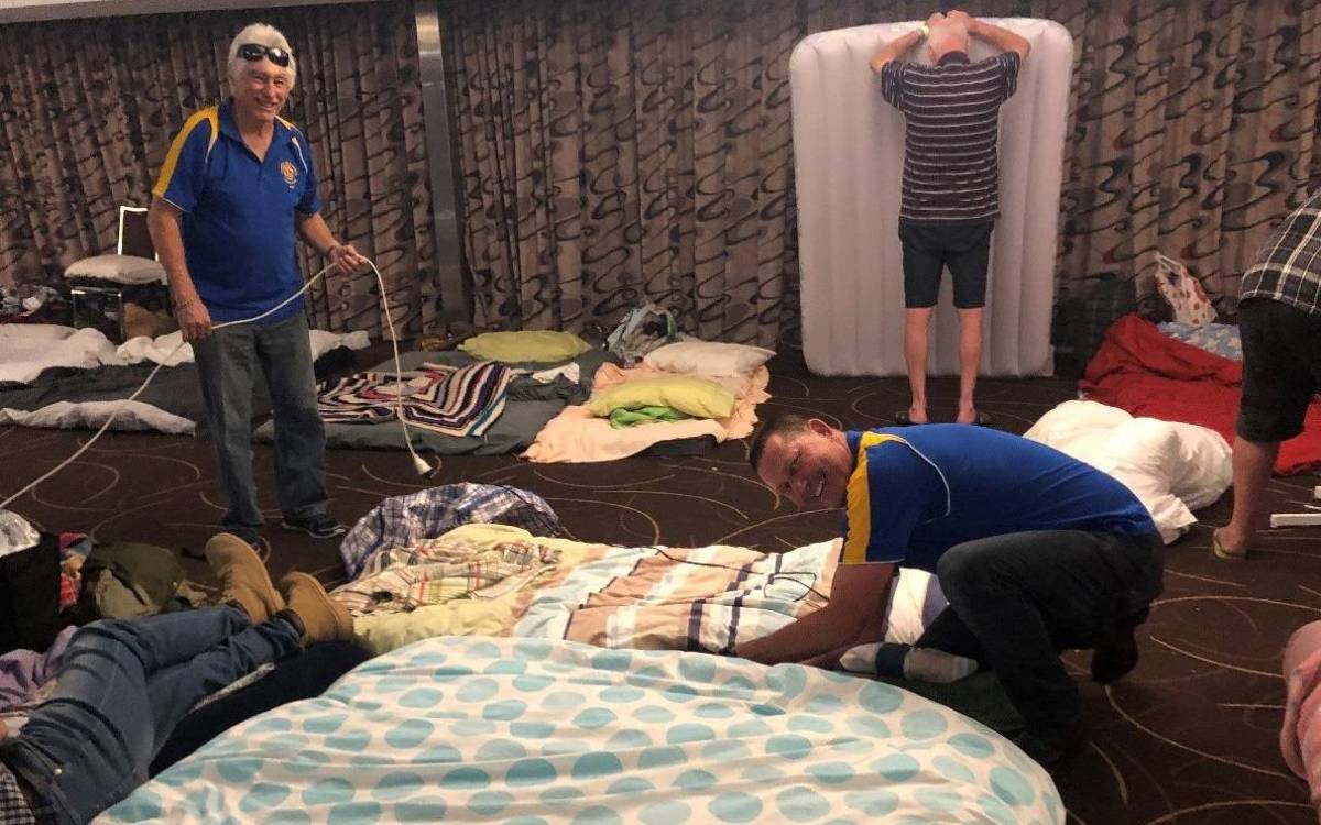 Two smiling men surrounded by air mattresses and bedding in a large room.