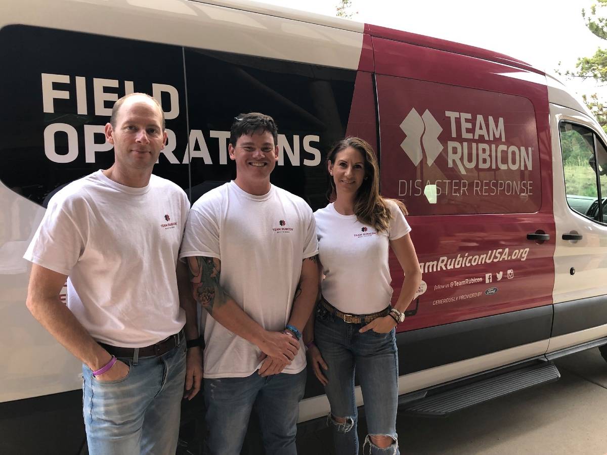 Three people pose for a camera in front of a van that says Team Rubicon Field Operations.