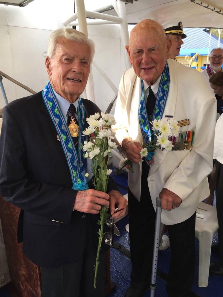 Two elderly men on board a ship. Both are carrying flowers and wearing medals.