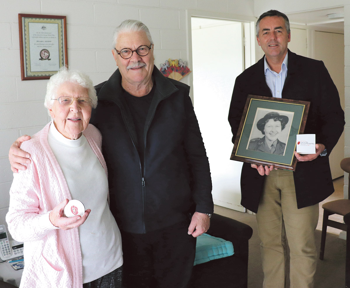 Middle-aged man poses inside a house next to elderly woman holding a medallion, and her son.