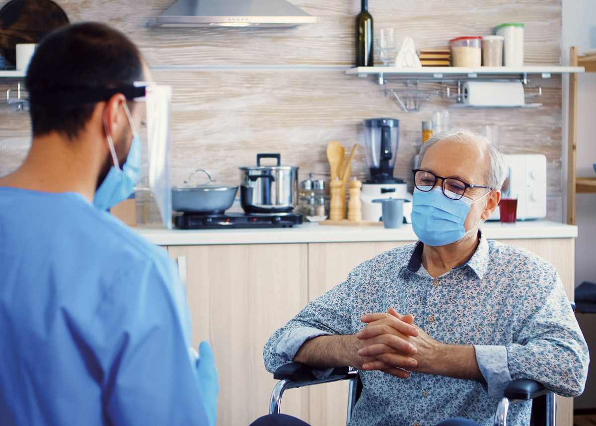 A male doctor standing up and a male patient sitting down in a chair. They are having a conversation whilst wearing face masks.