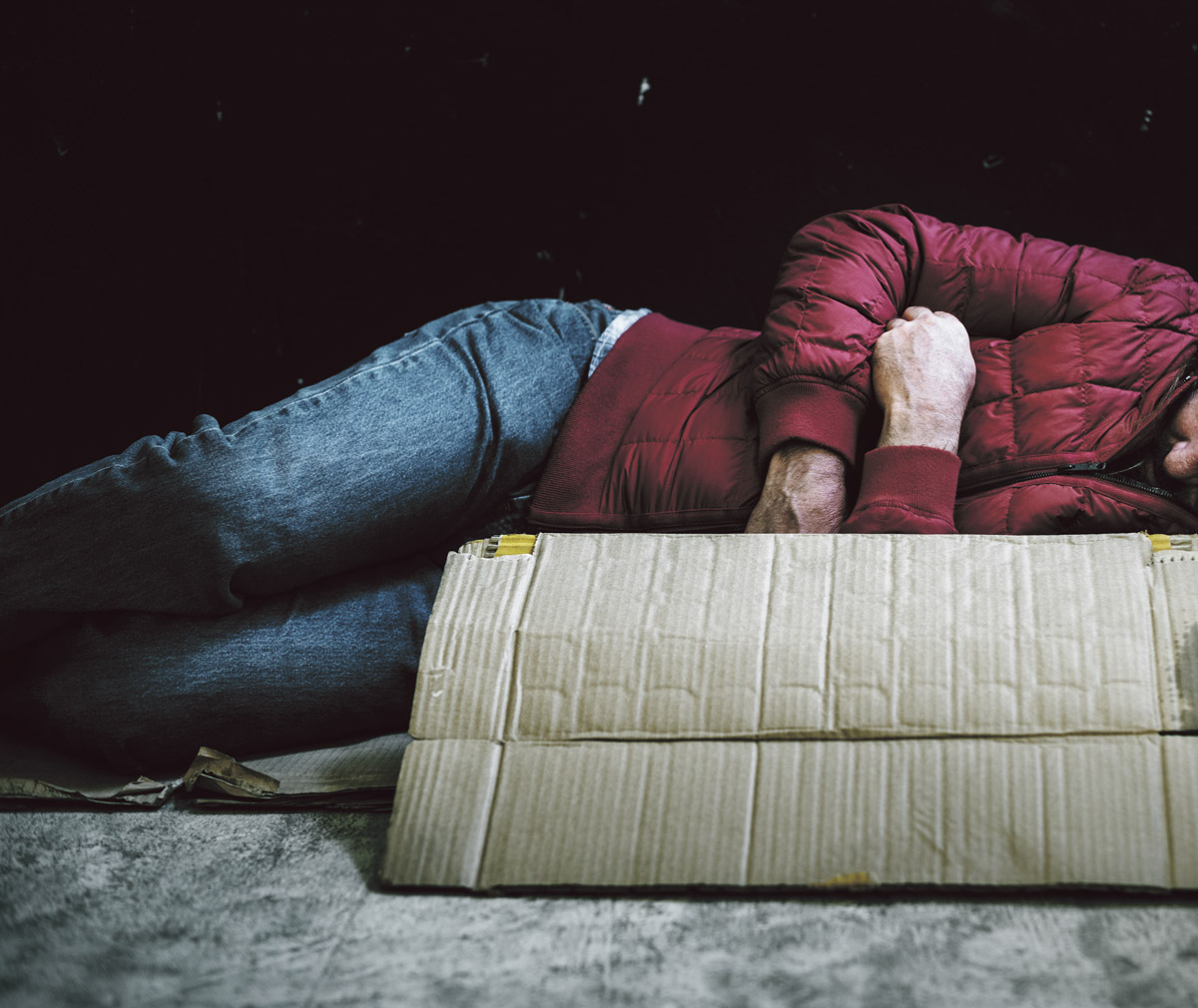 A homeless man sleeping rough with his arms folded.. He is wearing blue jeans and a red puffer jacket.