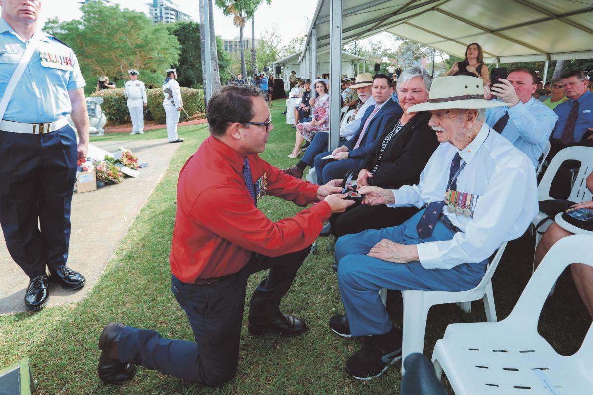 A young man, kneeling down on one knee, presents a medallion to an elderly veteran man who is sitting down on a plastic chair.