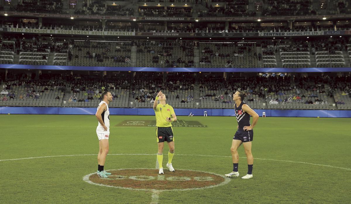 The AFL umpire throws a coin up into the air while two opposing AFL players look on. They are all standing in the middle of an oval surrounded by a large stadium.