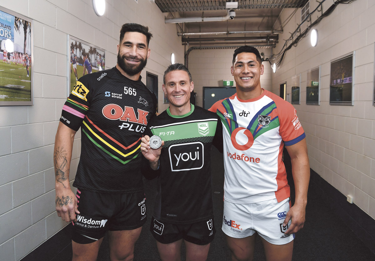 Three rugby league players, posing with the commemorative medallion and standing in a corridor.
