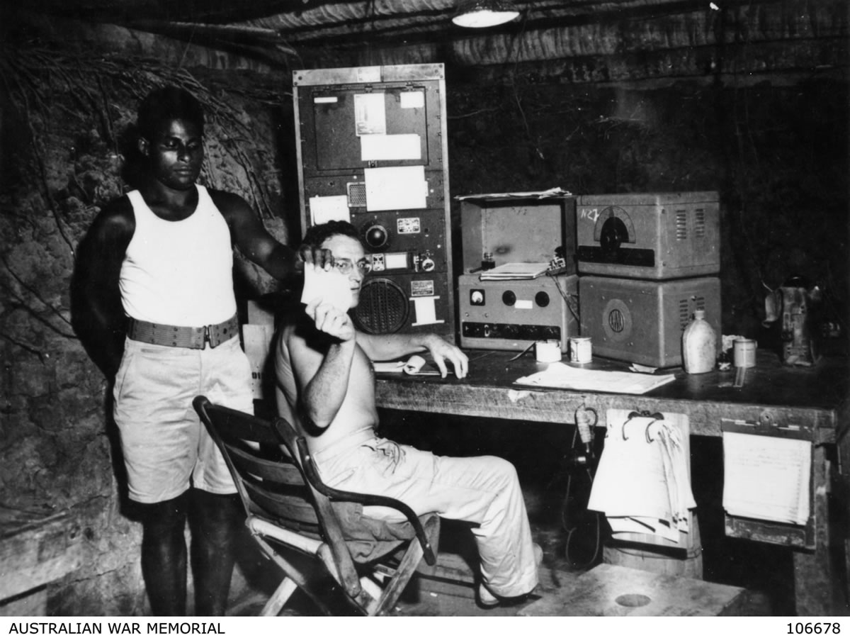 An Islander man standing and James Burrowes, an Australian coastwatcher, sitting down in a base radio station dugout in the Solomon Islands during World War Two.