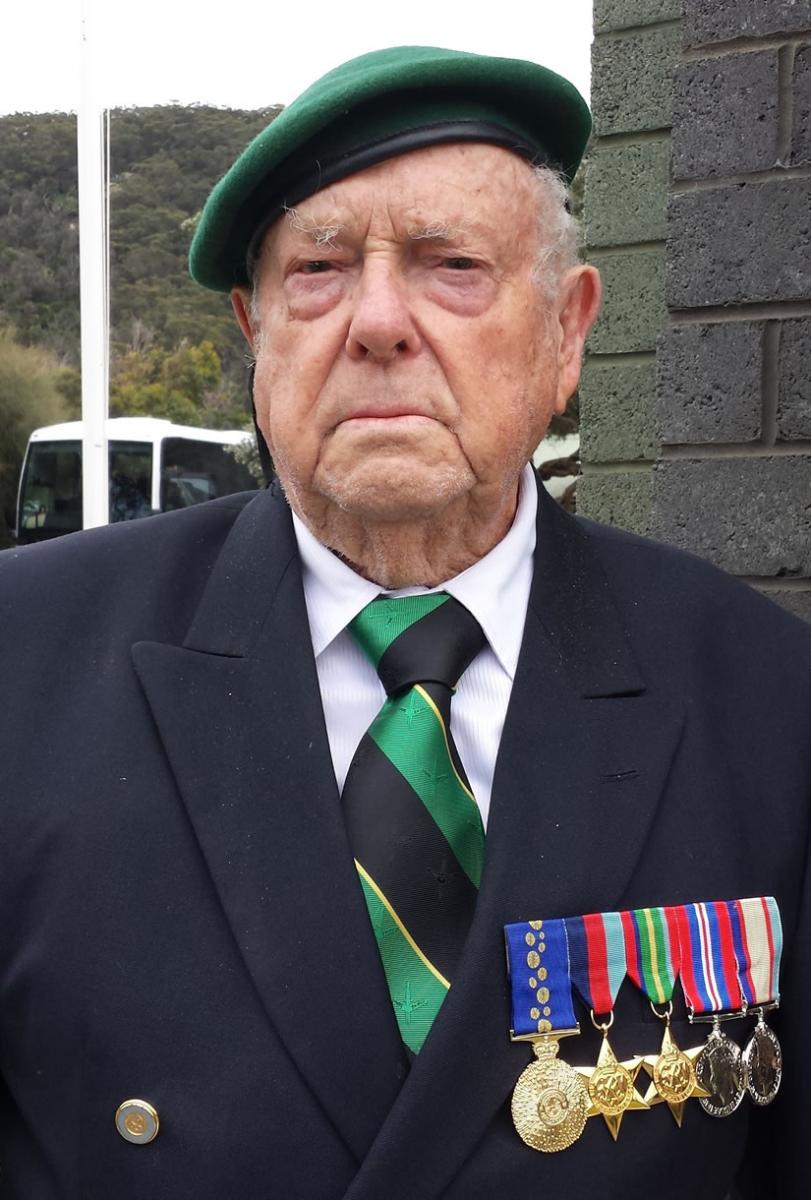A World War Two veteran man named James Burrowes. He is wearing a dark suit, green beret and a green and black tie with gold accents. Five service medals are pinned on the left of his chest.