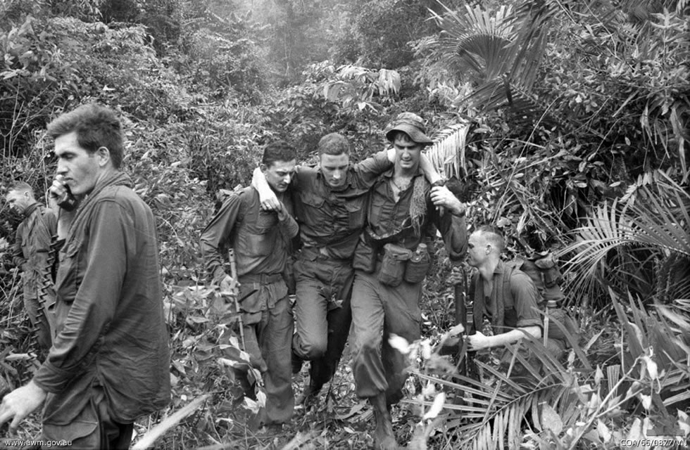 Australian soldiers in the jungle in October 1966 during the Vietnam War. One soldier is injured and is being supported by two other soldiers as he walks. Another soldier is on the radio requesting help.