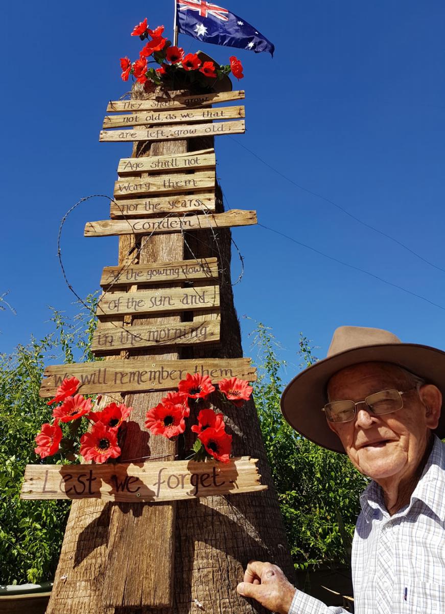 An elderly World War Two veteran man named Syd Kinsman alongside his home-made Anzac Day 2020 tribute. The tribute is fixed to a tree trunk. There are red poppies at Syd's eye level, an excerpt from the poem ‘For the Fallen’ by Laurence Binyon and an Australian flag and another bunch of red poppies on top.