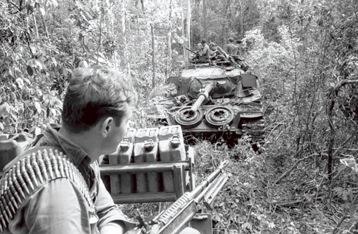 Black and white photo of a young man in uniform looking at a tank as they crash through jungle