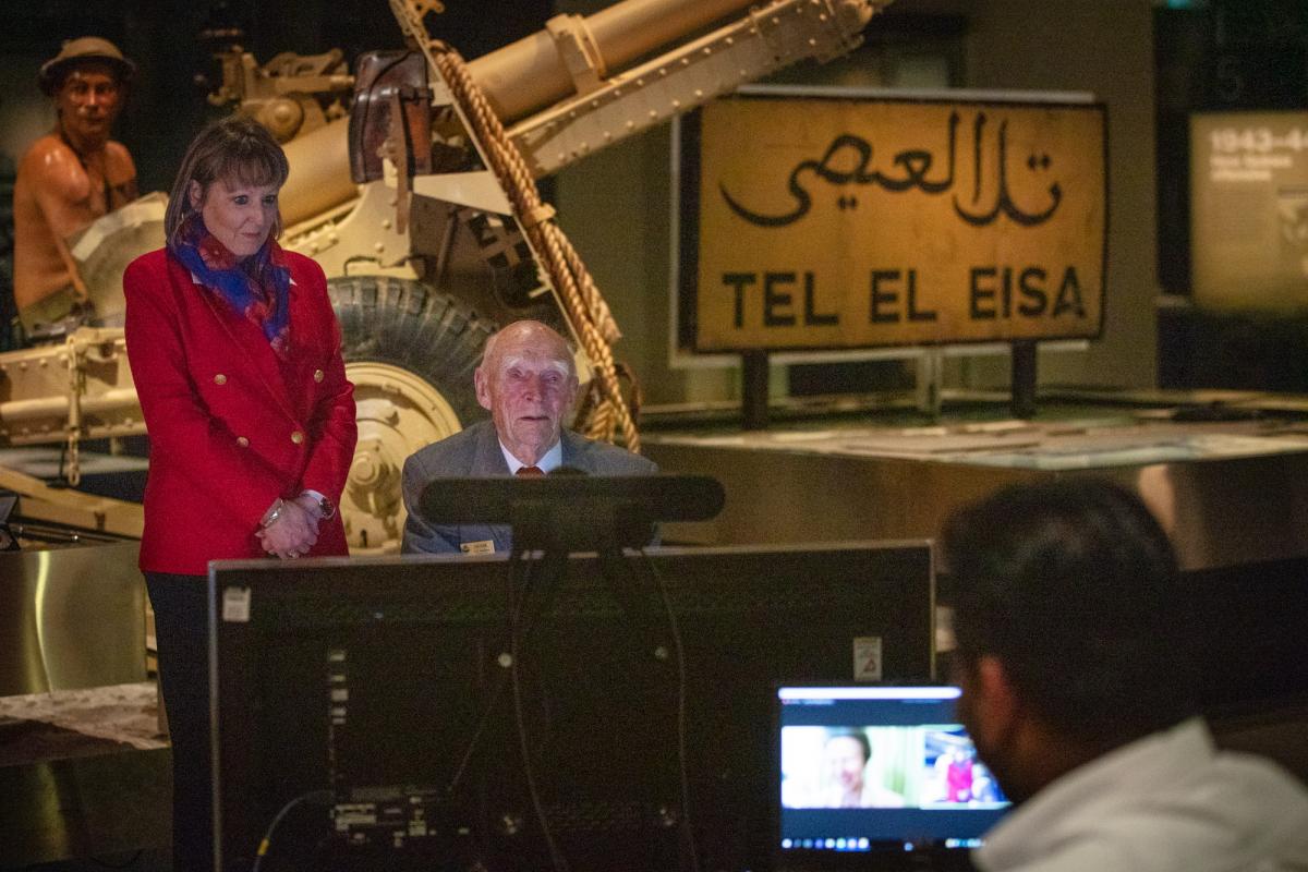 Elderly veteran sits talking to TV monitor with woman standing by his side and WWII display in background.