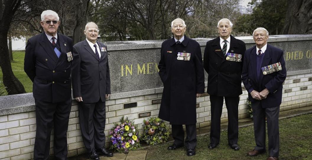 Five elderly men wearing medals stand in front of low wall with brass plaques attached