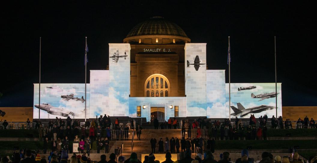 Night shot of front of AWM with crowd in foreground and various historic planes projected on the facade.
