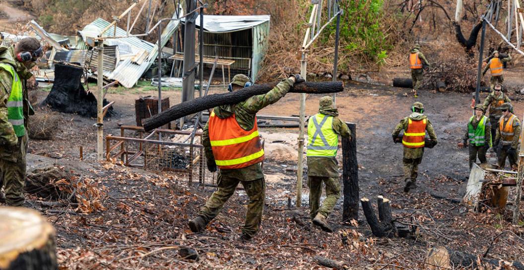 Australian Defence Force (ADF) reservists wearing high-visibility vests over their camouflage uniform. They are removing and carrying branches, logs and stumps away from the bushforest. There is the burnt remains of a large shed or house and a small shed.