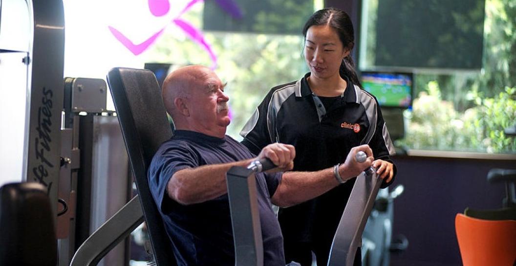 Young woman watches over elderly man on weights machine in gym