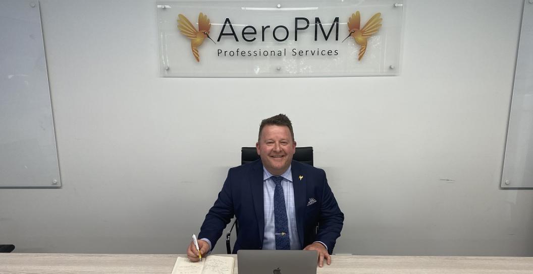 Smiling middle-aged man in office with sign reading AeroPM