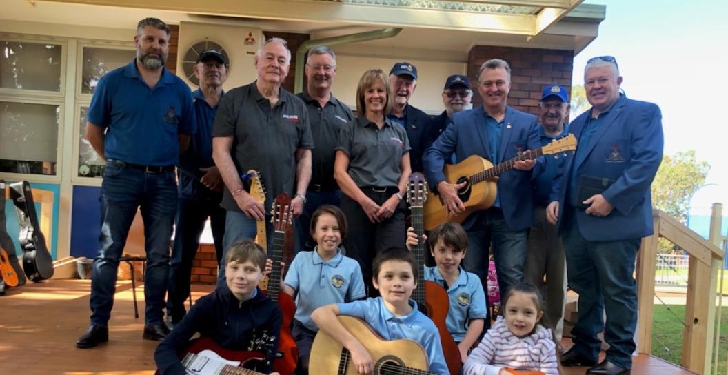 Several middle-aged people standing on an outside deck, with five children at their feet clutching guitars.