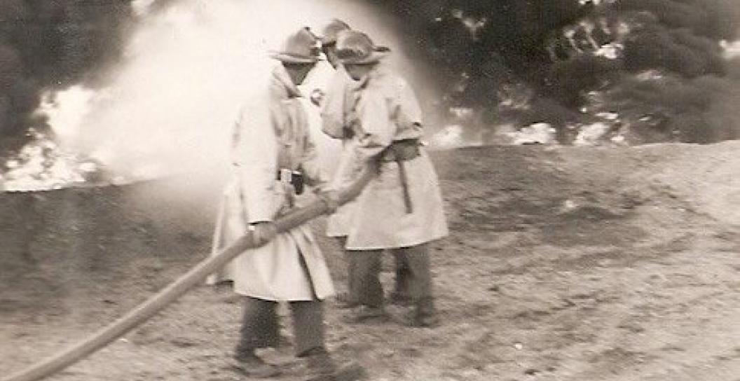 Black and white image of three ADF firefighters holding a fire hose with water spraying onto a fire