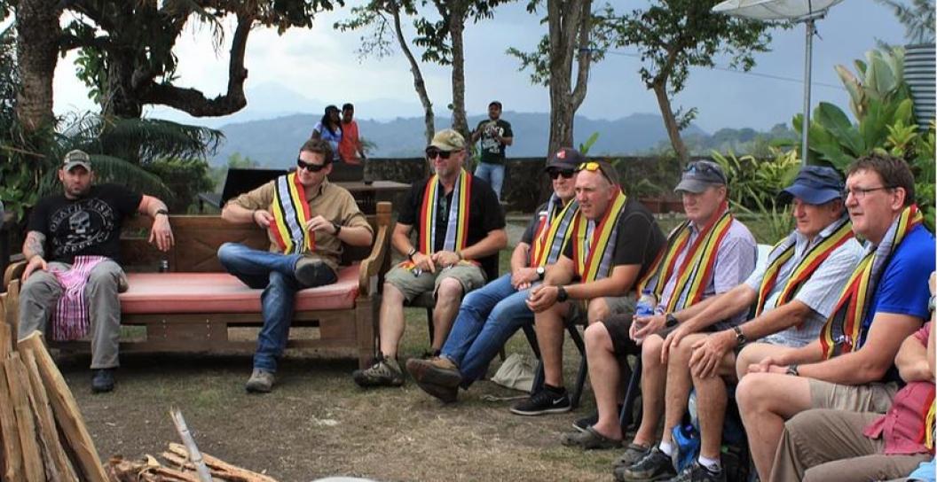 Eight or so men outside sitting in semi-circle listening to someone with tropical scenery in background