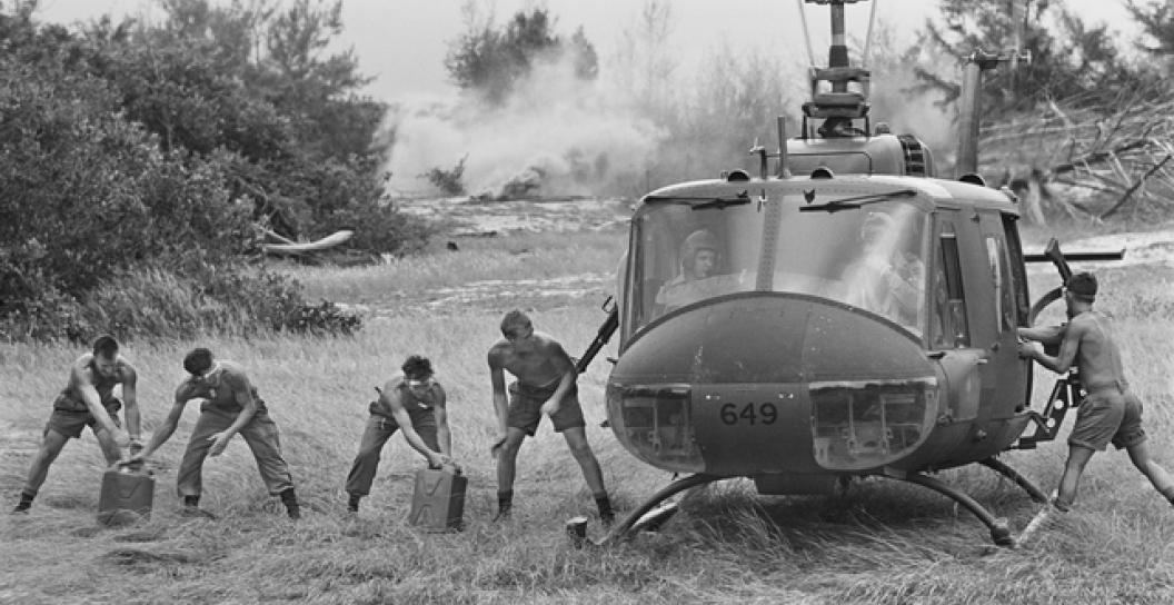 Five young soldiers unloading gerry cans from a Huey helicopter on an expanse of grass with bush in the background
