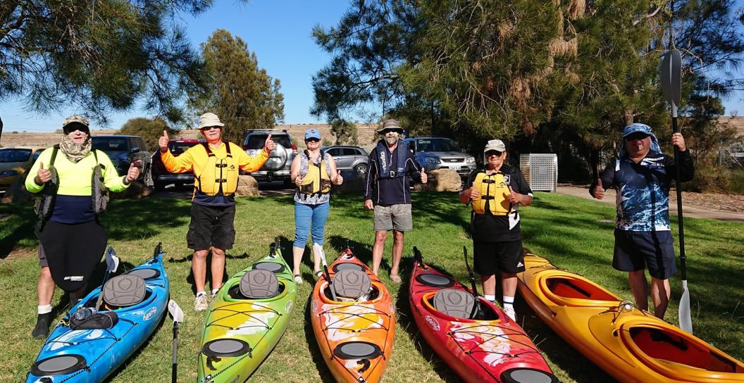 Six people pose next to their kayaks with thumbs up