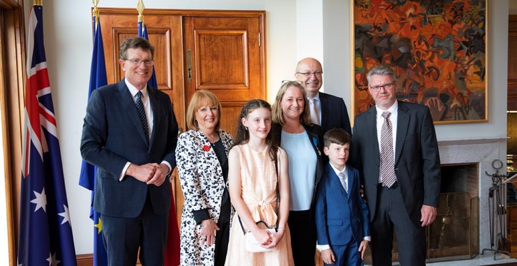 Three men, two women and two children pose for camera in large nicely decorated room, next to Australian flag