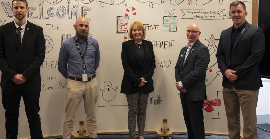 Five people are standing side by side facing the camera, all smiling. They are standing in front of a large white board.